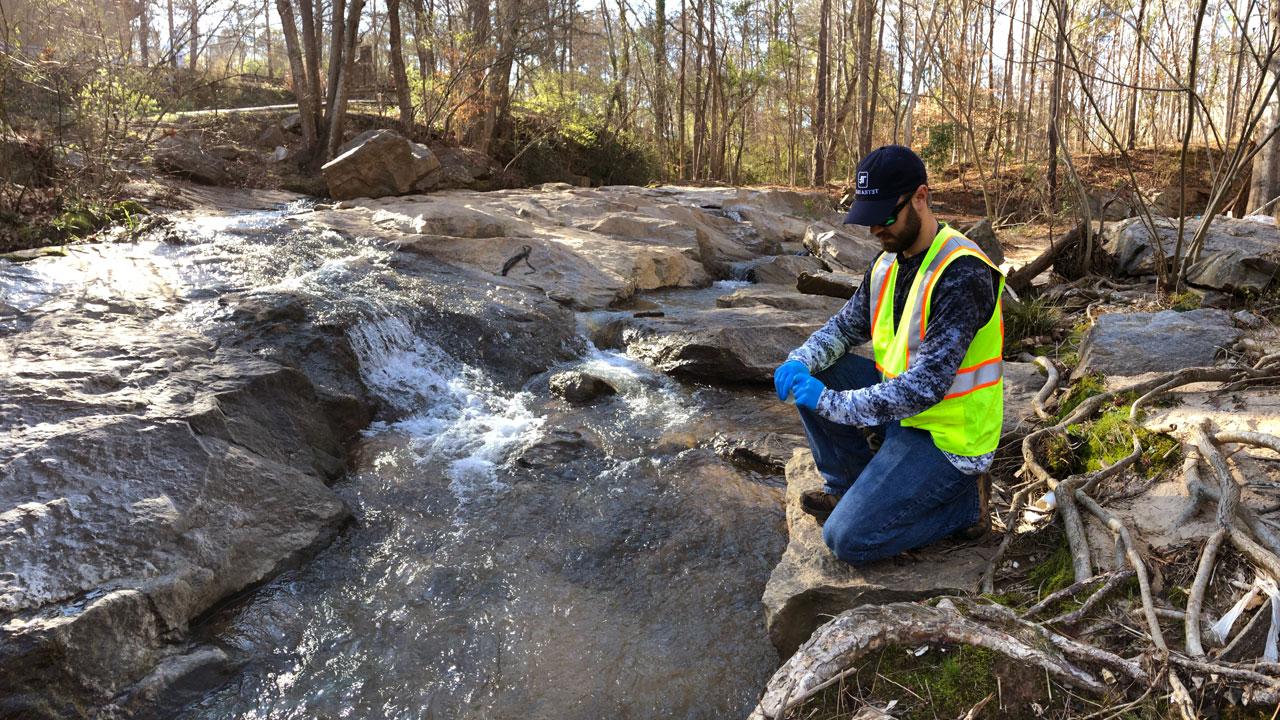Worker wearing safety gear kneeling on a rock preparing to collect a stream water sample